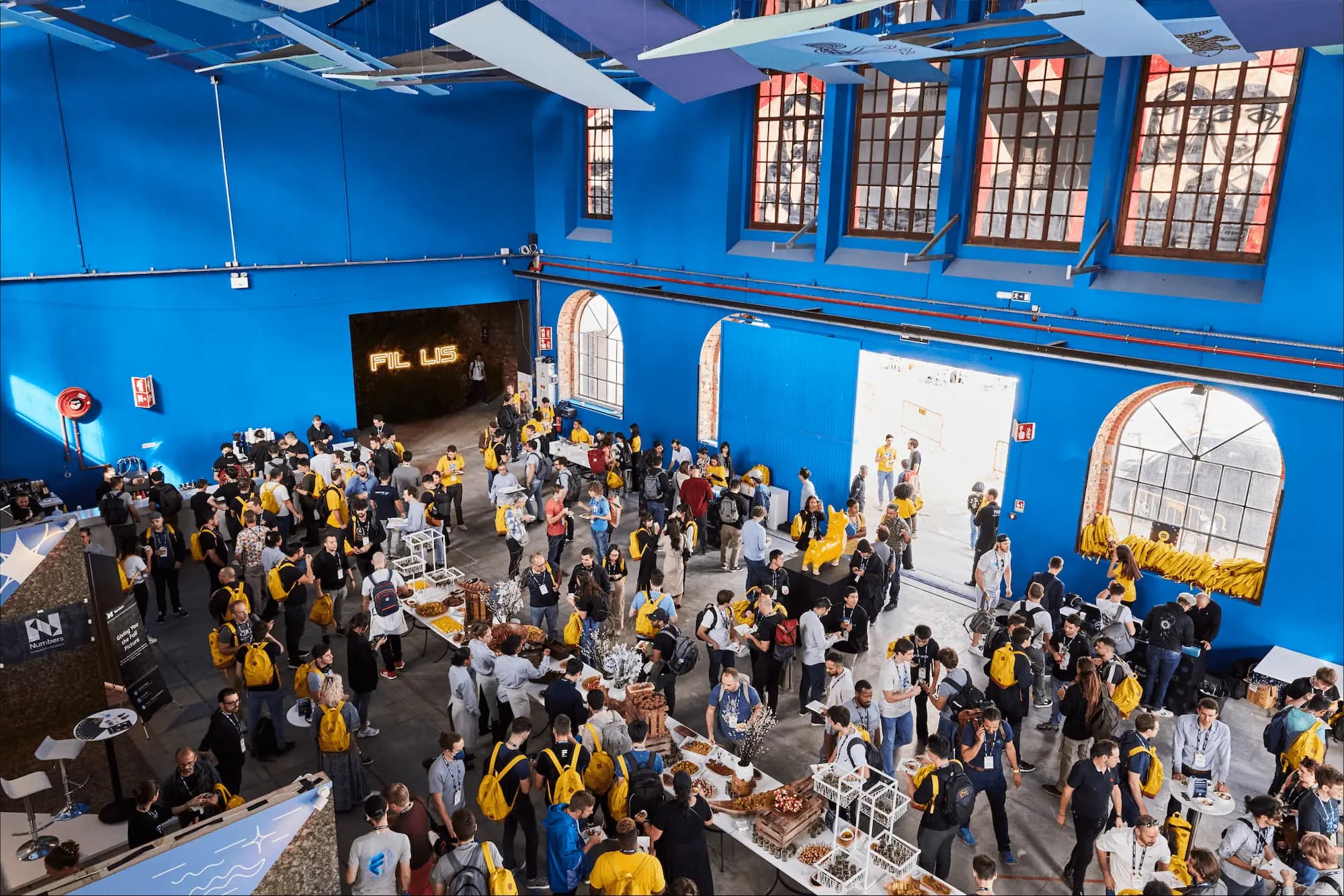 Large event space with people gathered around tables for networking, many wearing yellow backpacks, against a bright blue backdrop.