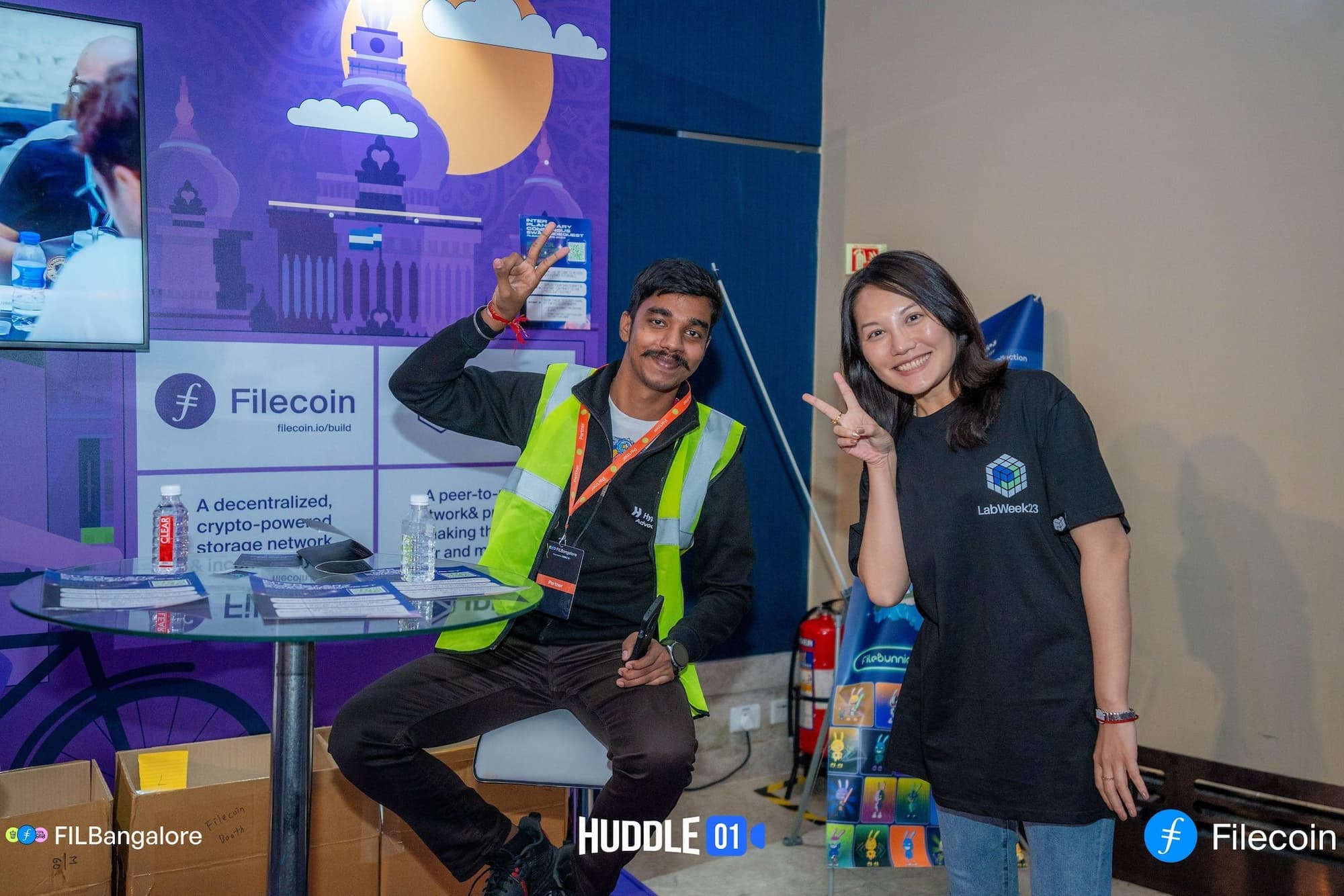 A man and a woman posing and making peace signs at a Filecoin event booth. The man is sitting and wearing a green high-visibility vest, while the woman is standing and wearing a black 'LabWeek23' t-shirt. The booth has a purple backdrop with Filecoin branding and information, including a depiction of a cityscape. Bottles of water and promotional materials are on the table in front of them.